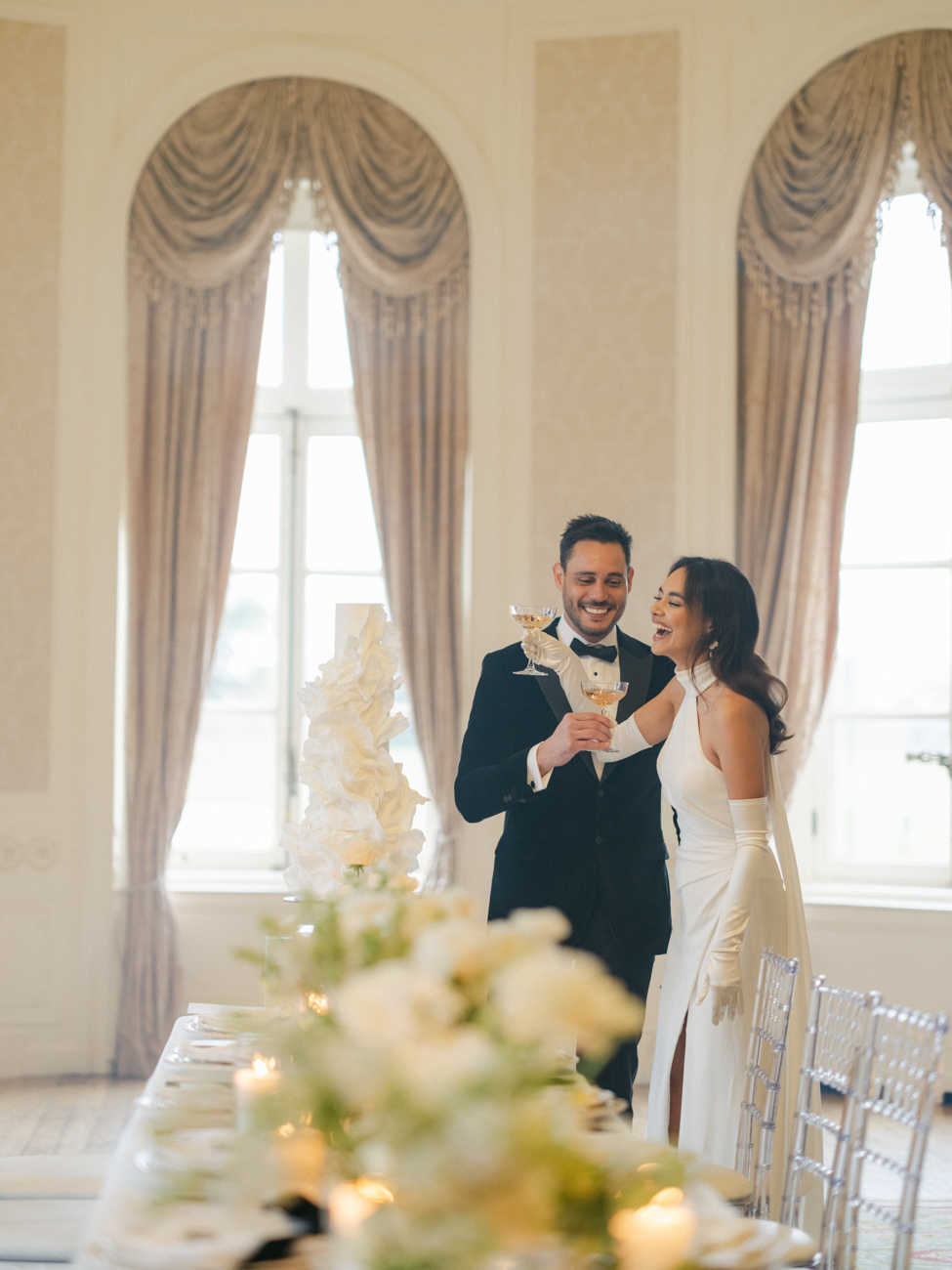 bride and groom laughing together next to luxury cake holding champagne