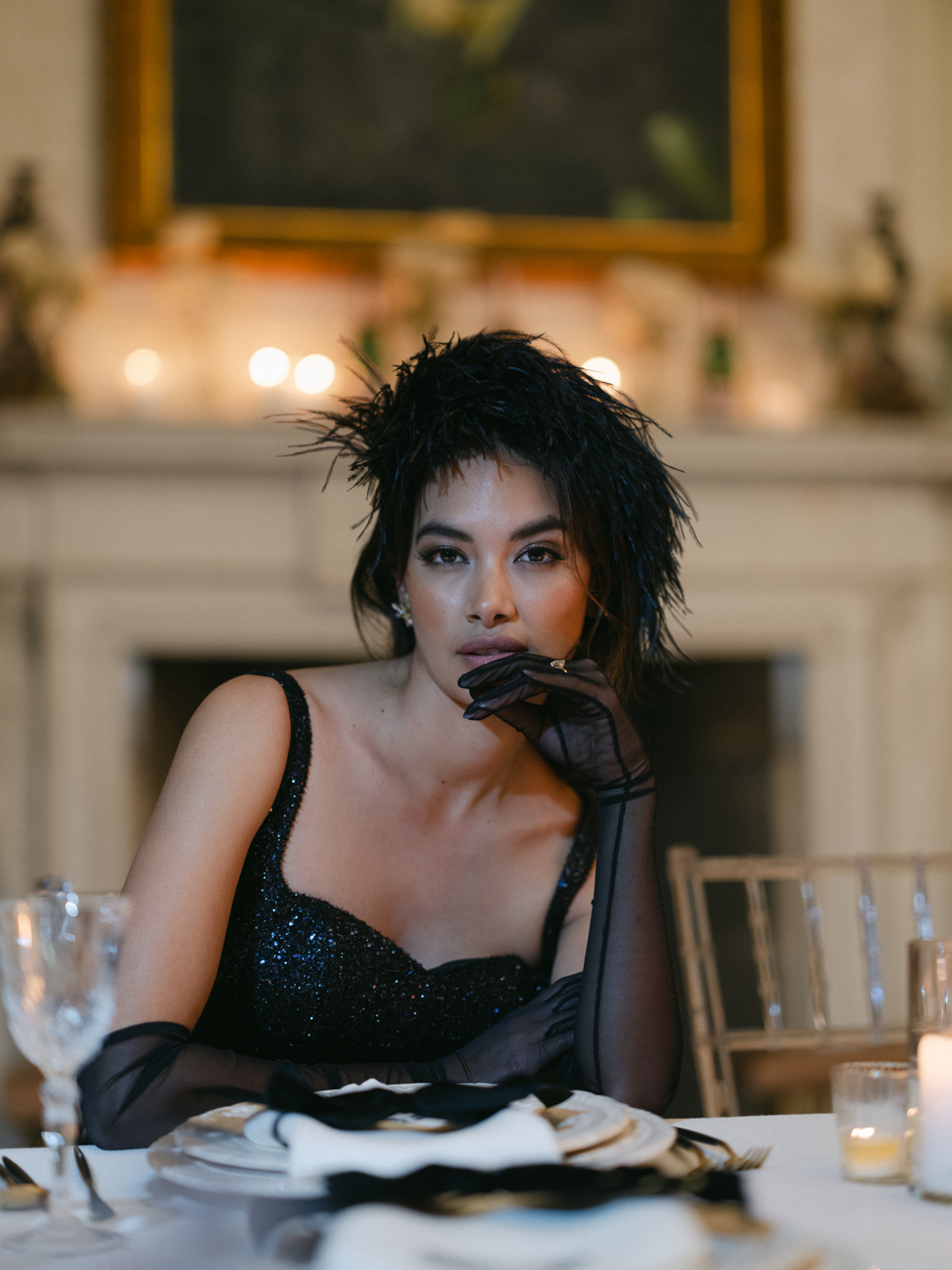 girl sitting at table during wedding reception with a Saltburn feel. Wearing black feather head piece and sheer black gloves